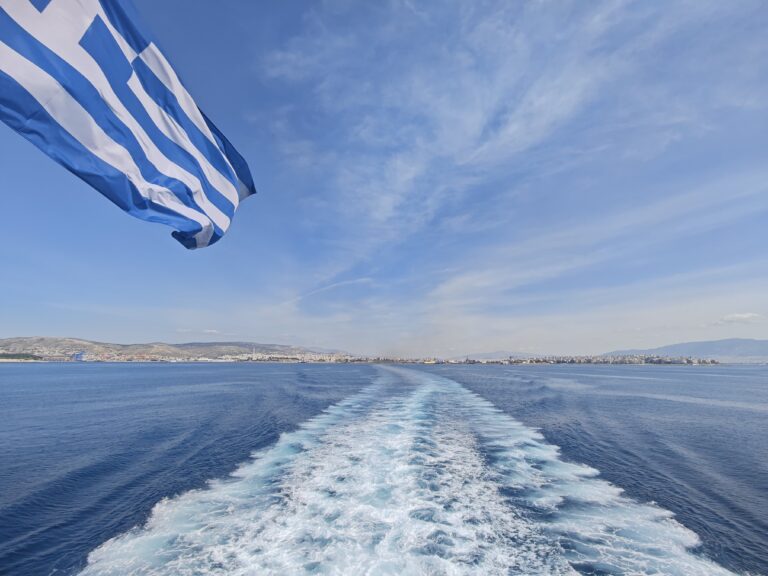 Greek flag waving above the sea as the ferry leaves a trail toward the coast