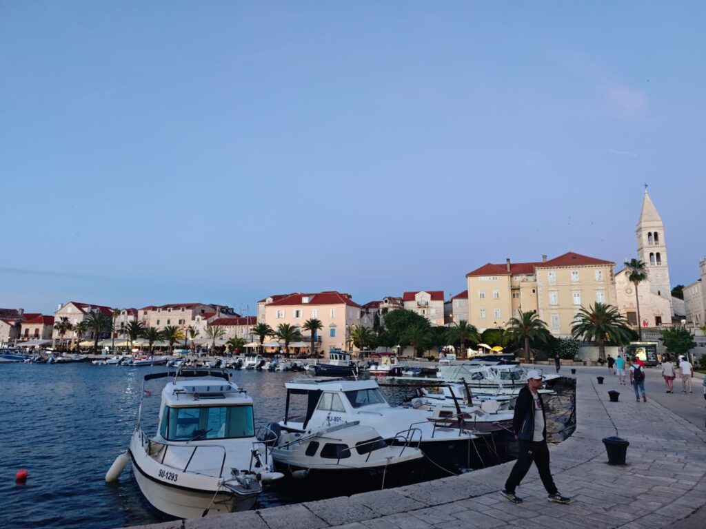 Evening view of Supetar harbour on Brač Island, with fishing boats, palm trees and historic stone buildings along the waterfront.