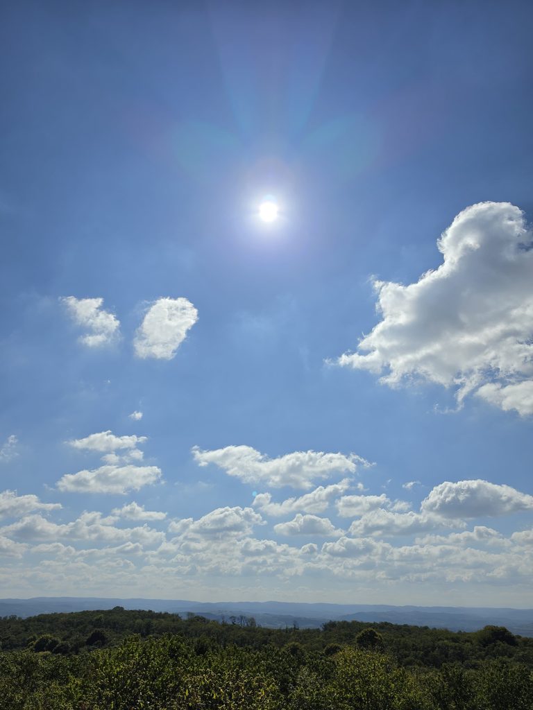 Za ovu fotografiju upiši u **Alt Text**:
**Sunce i bijeli oblaci nad šumovitim krajolikom Hrastovačke gore.**